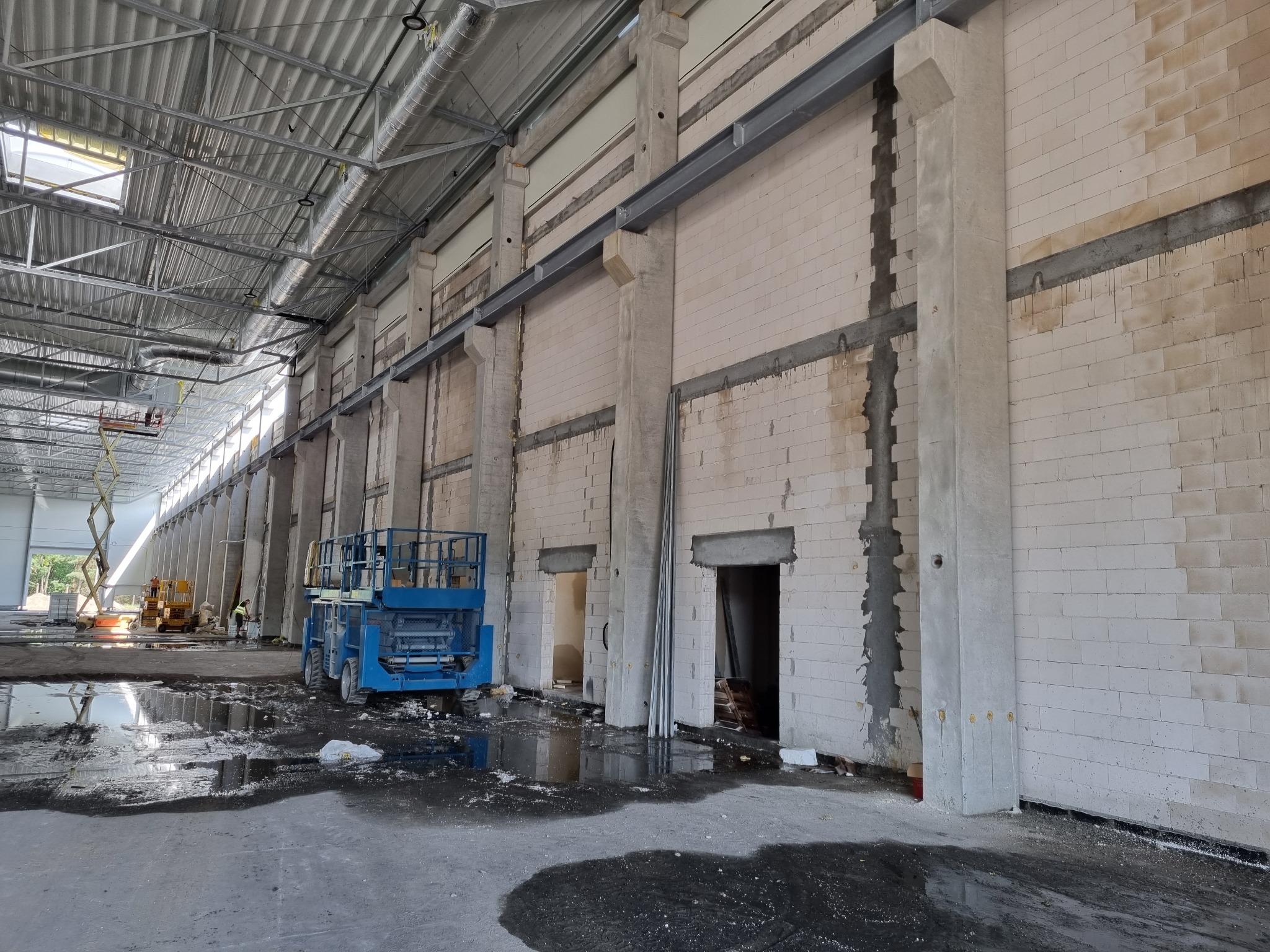 Wnętrze hali produkcyjnej w trakcie prac budowlanych, widoczne surowe ściany z betonu komórkowego, stalowe belki, podnośniki nożycowe i kałuże na posadzce.