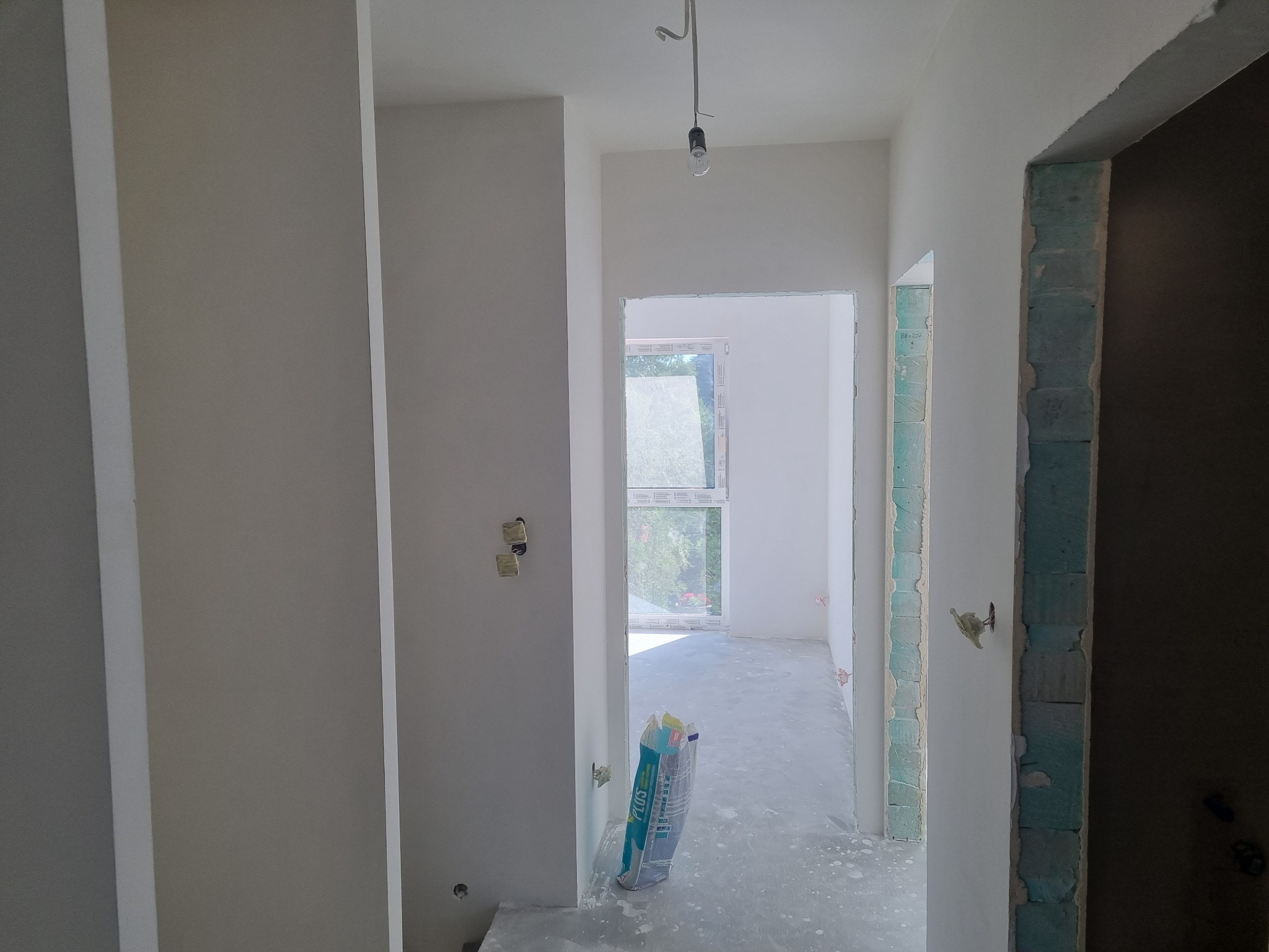 Wnętrze w trakcie prac wykończeniowych: świeżo otynkowane ściany, widoczny worek z zaprawą gipsową, surowa żarówka na kablu, przygotowane otwory drzwiowe z widocznym bloczkiem betonu komórkowego.