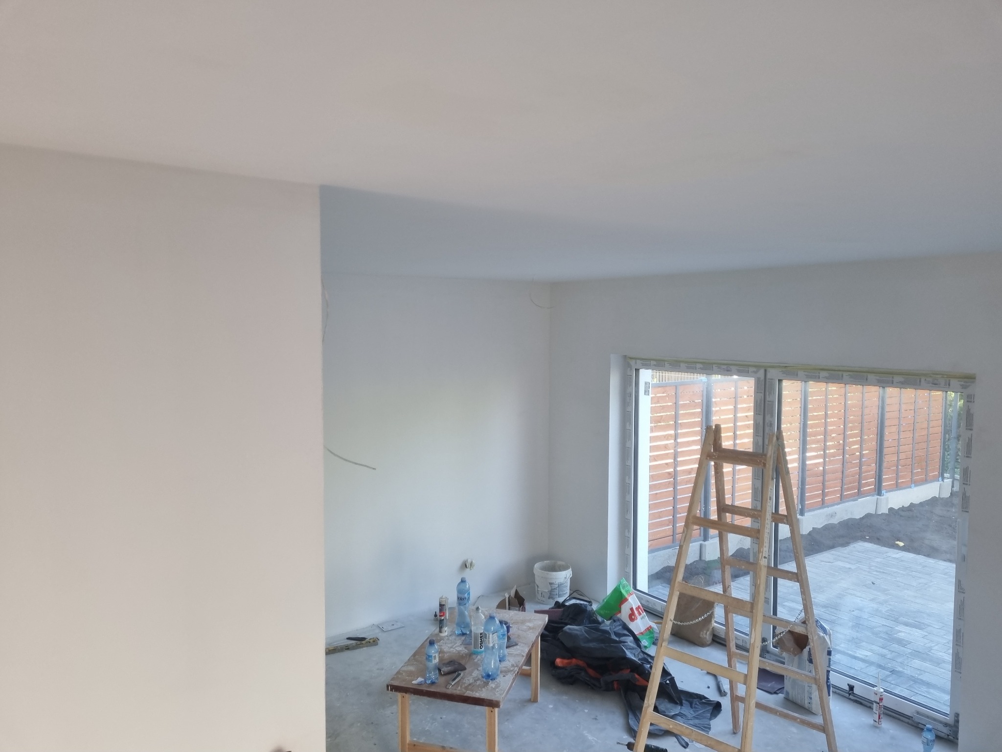 Wnętrze świeżo pomalowanego pokoju w Toruniu, widoczna drabina oparta o okno tarasowe, stolik z butelkami wody i narzędziami malarskimi, ściany i sufit w świeżej, białej farbie.