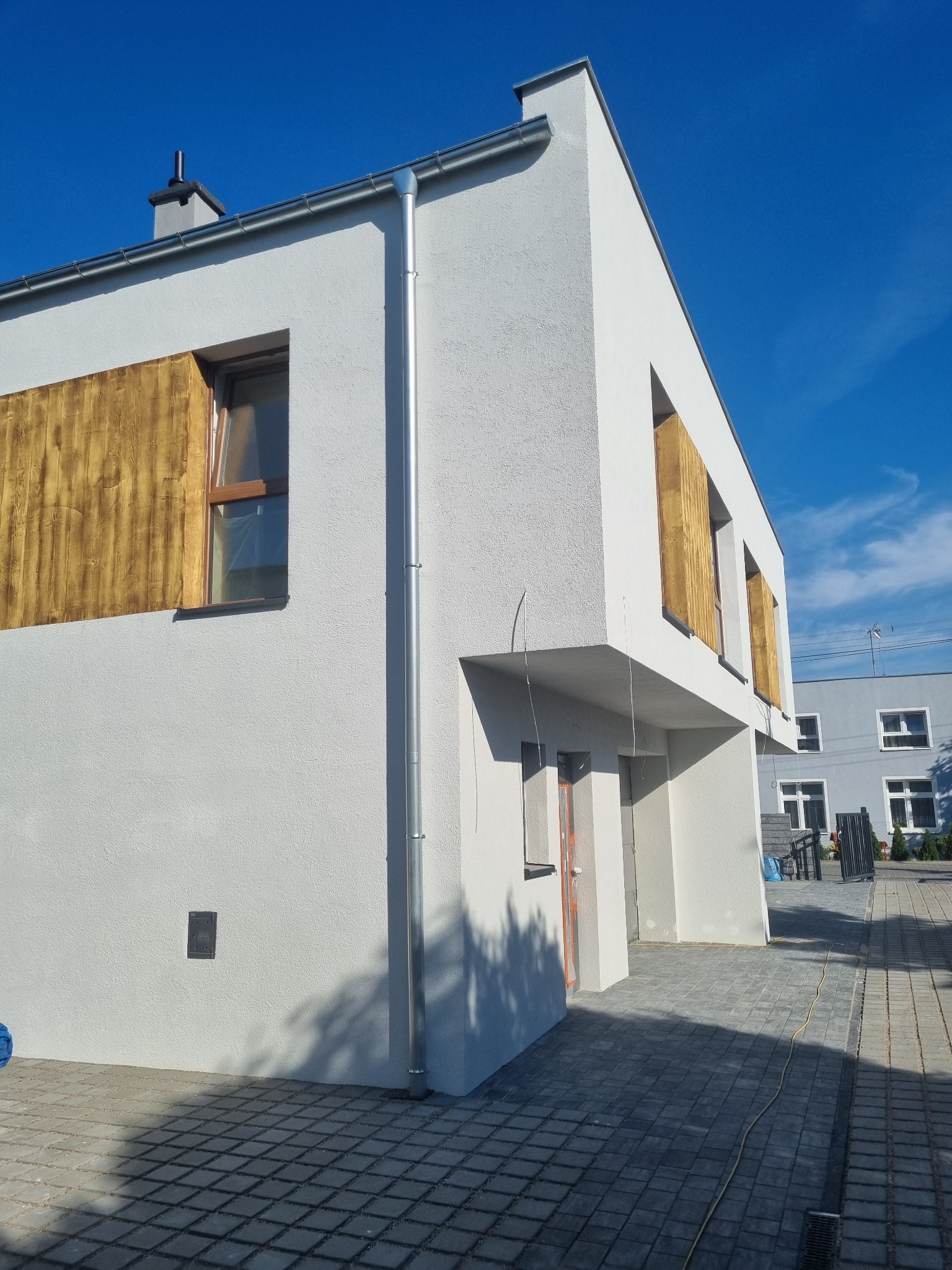 Nowoczesny dom z białą elewacją i drewnianymi akcentami wokół okien, widoczne orynnowanie i kostka brukowa na podwórku.