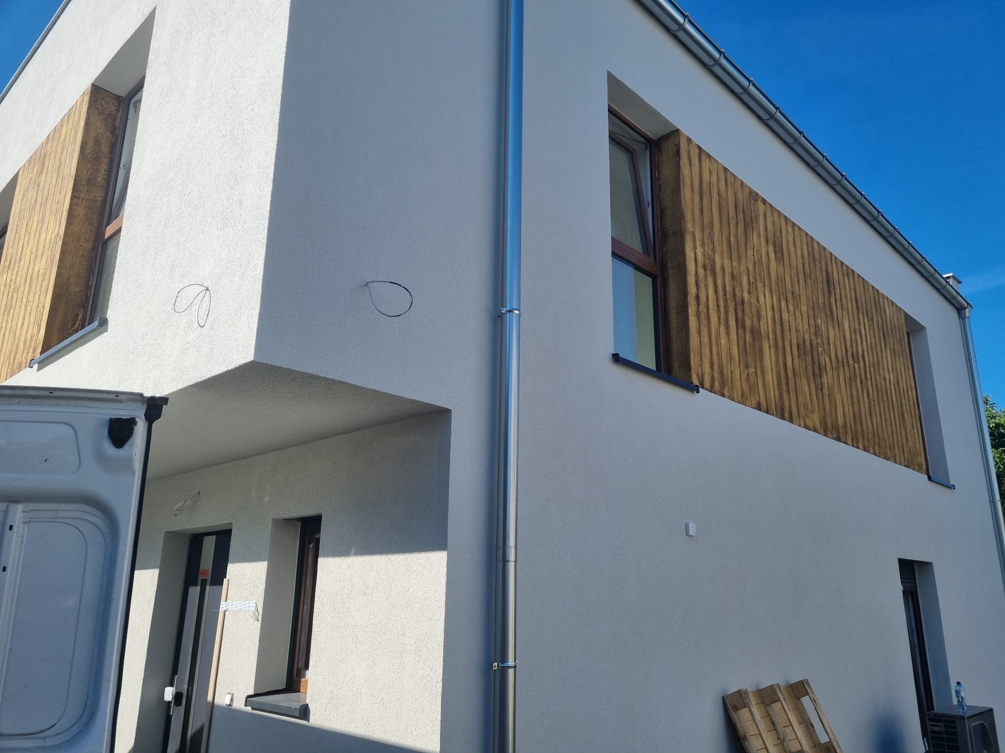 Nowoczesny dom z jasną elewacją, drewnianymi panelami dekoracyjnymi wokół okien i srebrną rynną, widoczna boczna ściana furgonetki.