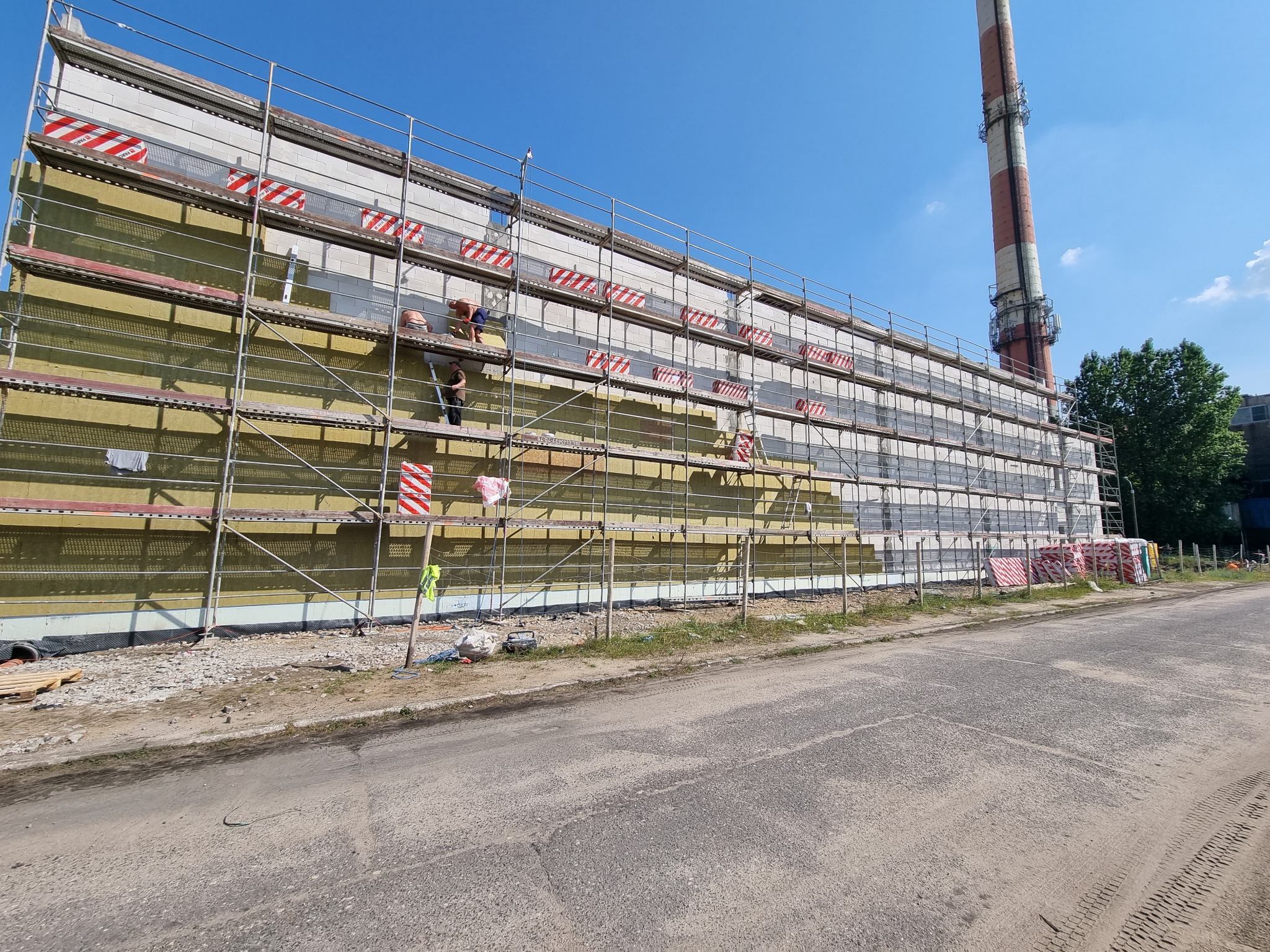 Prace przy ocieplaniu ściany budynku przemysłowego z wysokim kominem w tle, widoczne rusztowania i pracownicy montujący izolację termiczną.