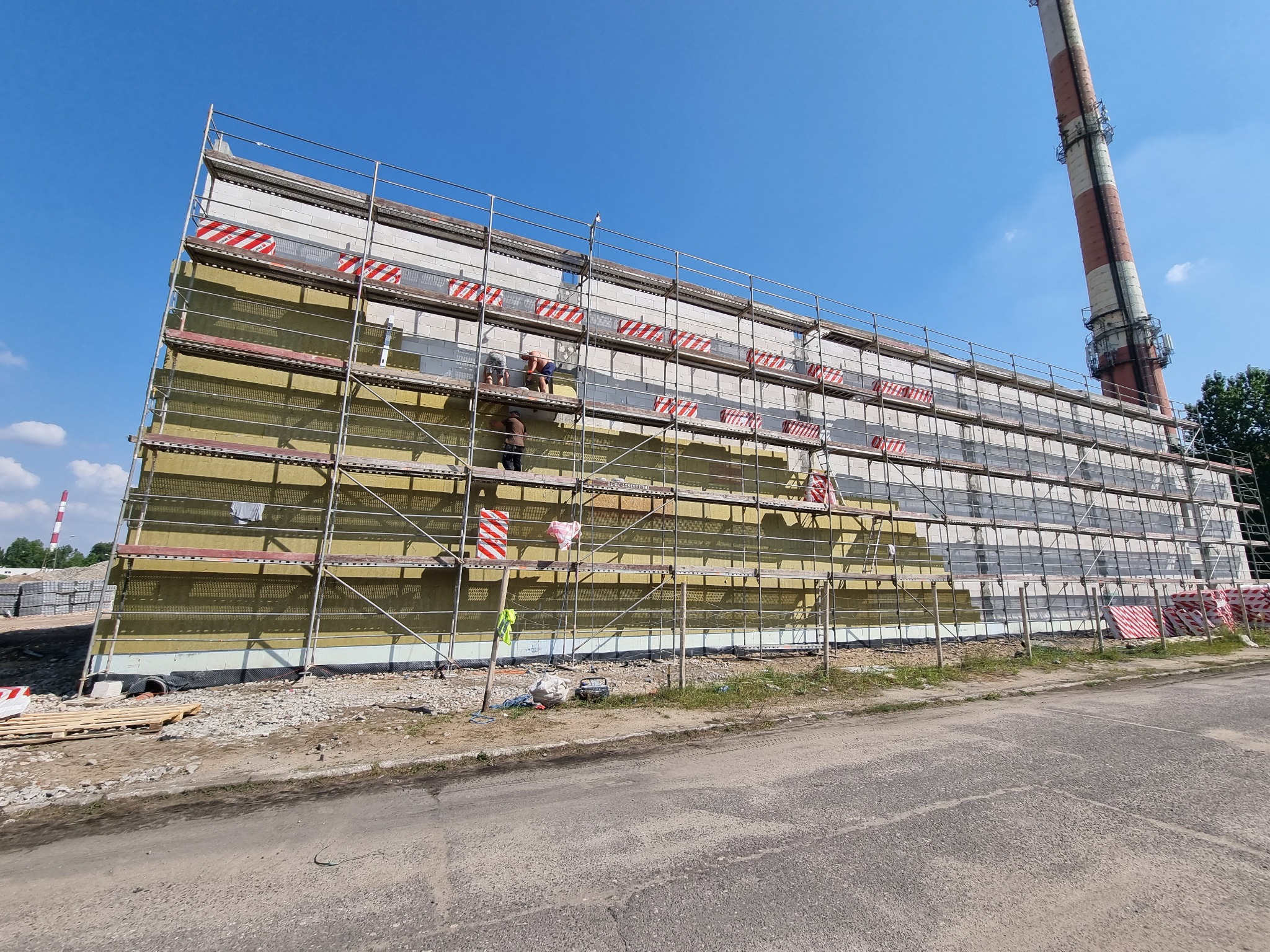 Ocieplanie elewacji budynku przemysłowego z widocznymi rusztowaniami, izolacją z wełny mineralnej i pracownikami w trakcie montażu, w tle komin przemysłowy.