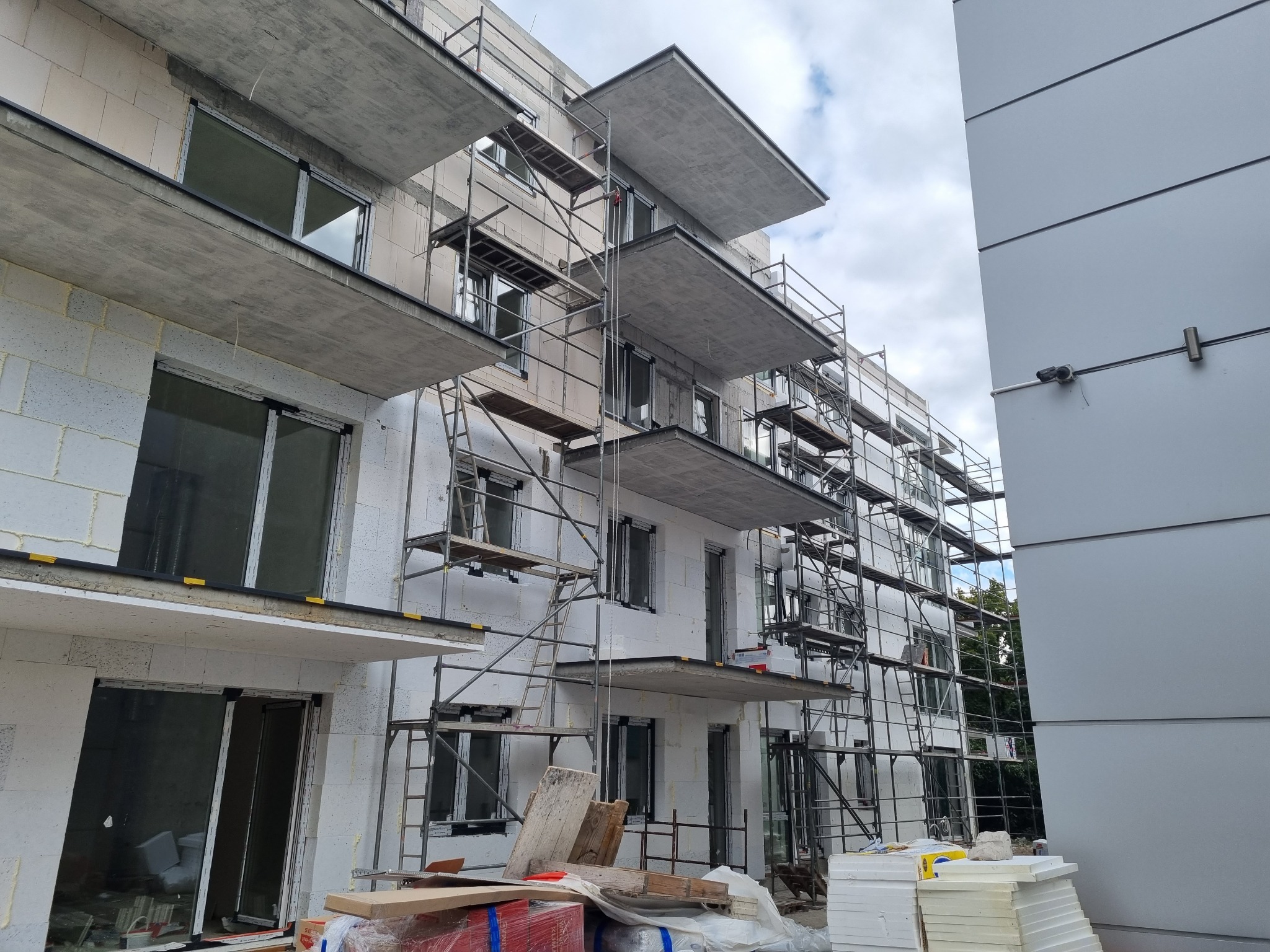 Budynek wielorodzinny w trakcie prac wykończeniowych elewacji, widoczne rusztowania, materiały budowlane i elementy izolacji na balkonach.