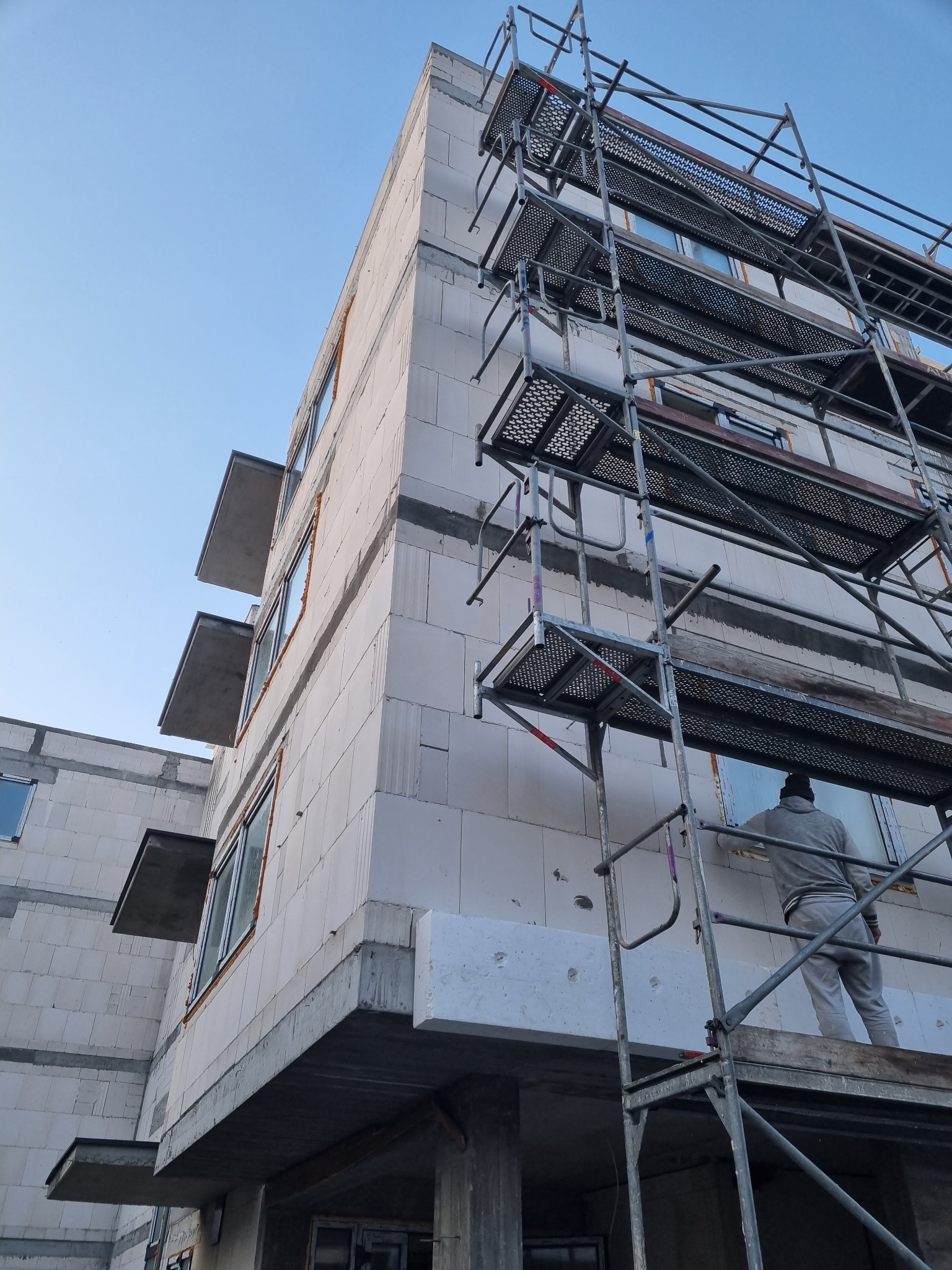Budynek wielorodzinny w trakcie prac wykończeniowych elewacji, widoczne rusztowania i pracownik w szarym kombinezonie montujący izolację termiczną na ścianie z betonu komórkowego.