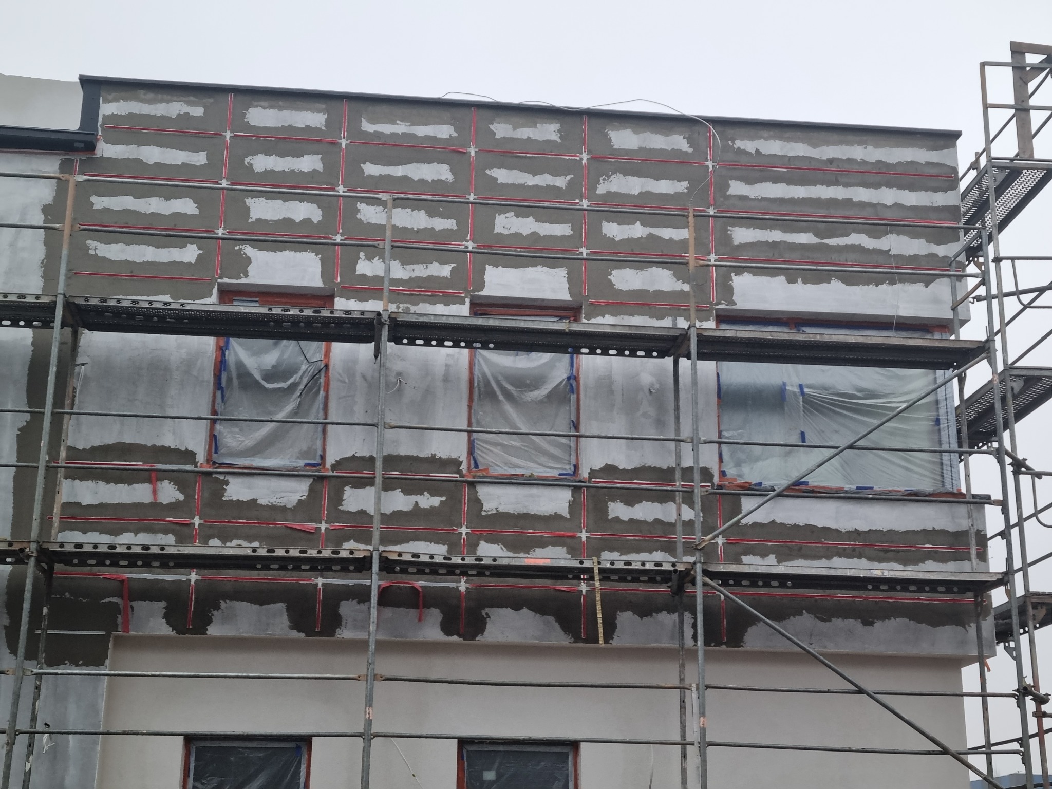 Elewacja budynku w trakcie przygotowania do ocieplenia, widoczne rusztowania, okna zabezpieczone folią, ściany z nałożoną warstwą bazową z siatką wzmacniającą.