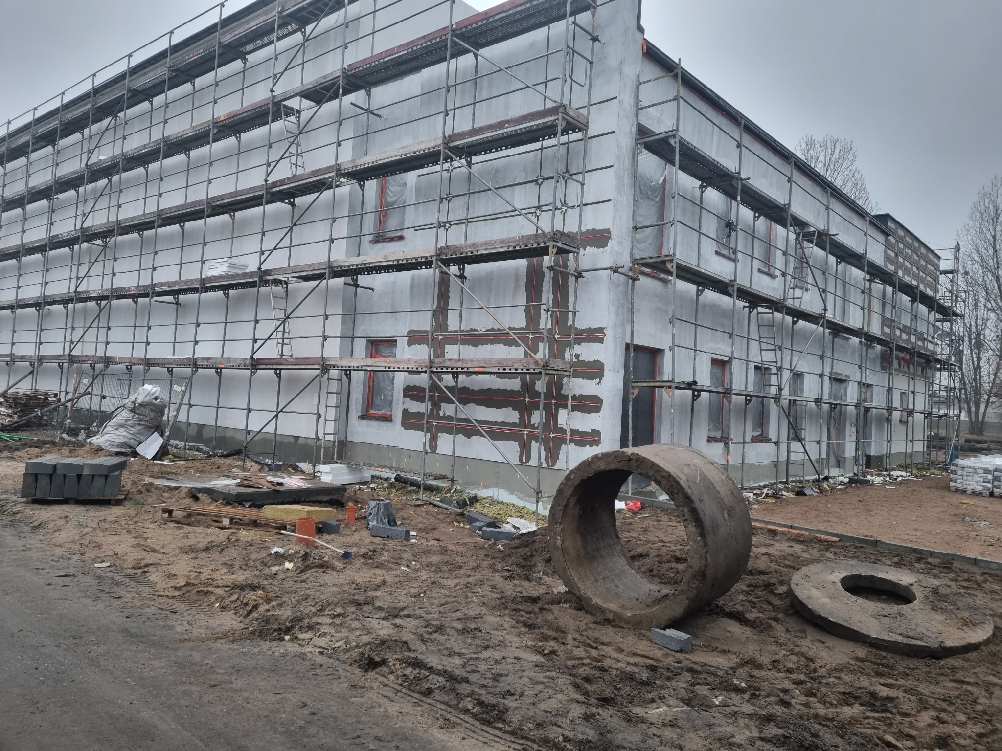 Budynek w trakcie prac elewacyjnych, otoczony rusztowaniami, z widocznymi elementami izolacji i betonowymi kręgami na pierwszym planie, na zewnątrz budynku.