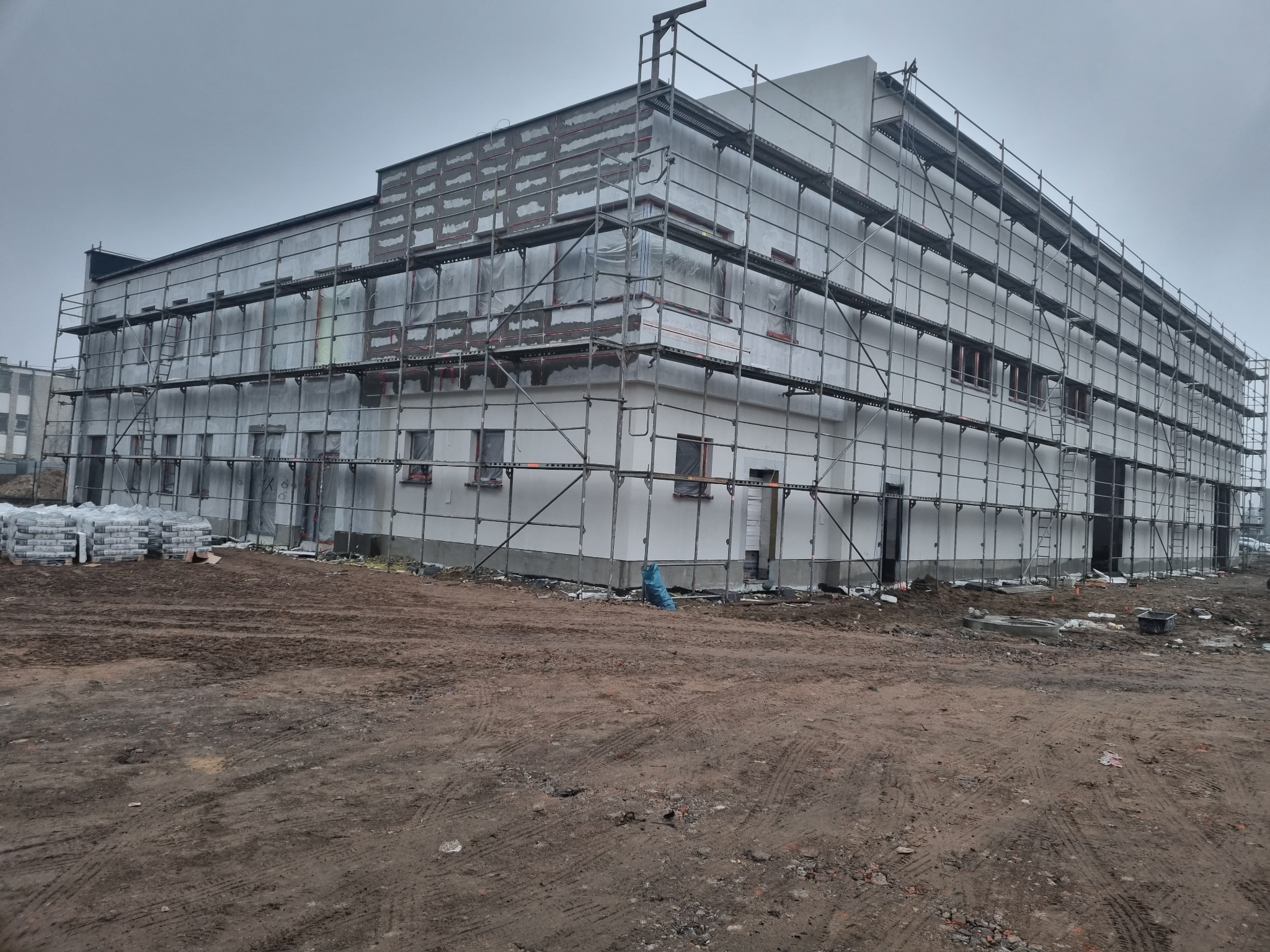 Budynek w trakcie prac elewacyjnych, otoczony rusztowaniami, z widocznymi workami materiałów budowlanych i błotnistym terenem wokół.