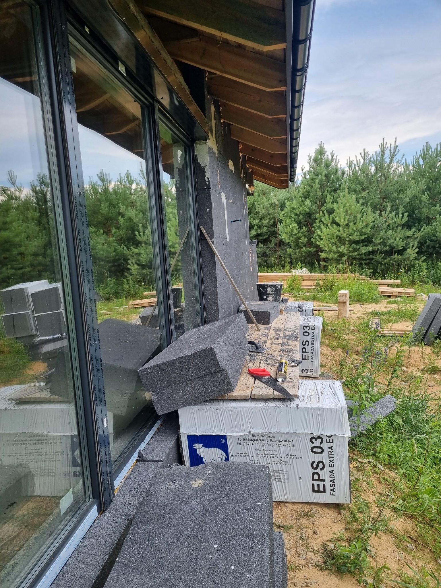 Fragment elewacji budynku w trakcie ocieplania, widoczne płyty styropianowe EPS 03, przycięte i częściowo zamontowane, narzędzia pracy na palecie, duże okno z profilami Alufrof, tło stanowią drzewa.