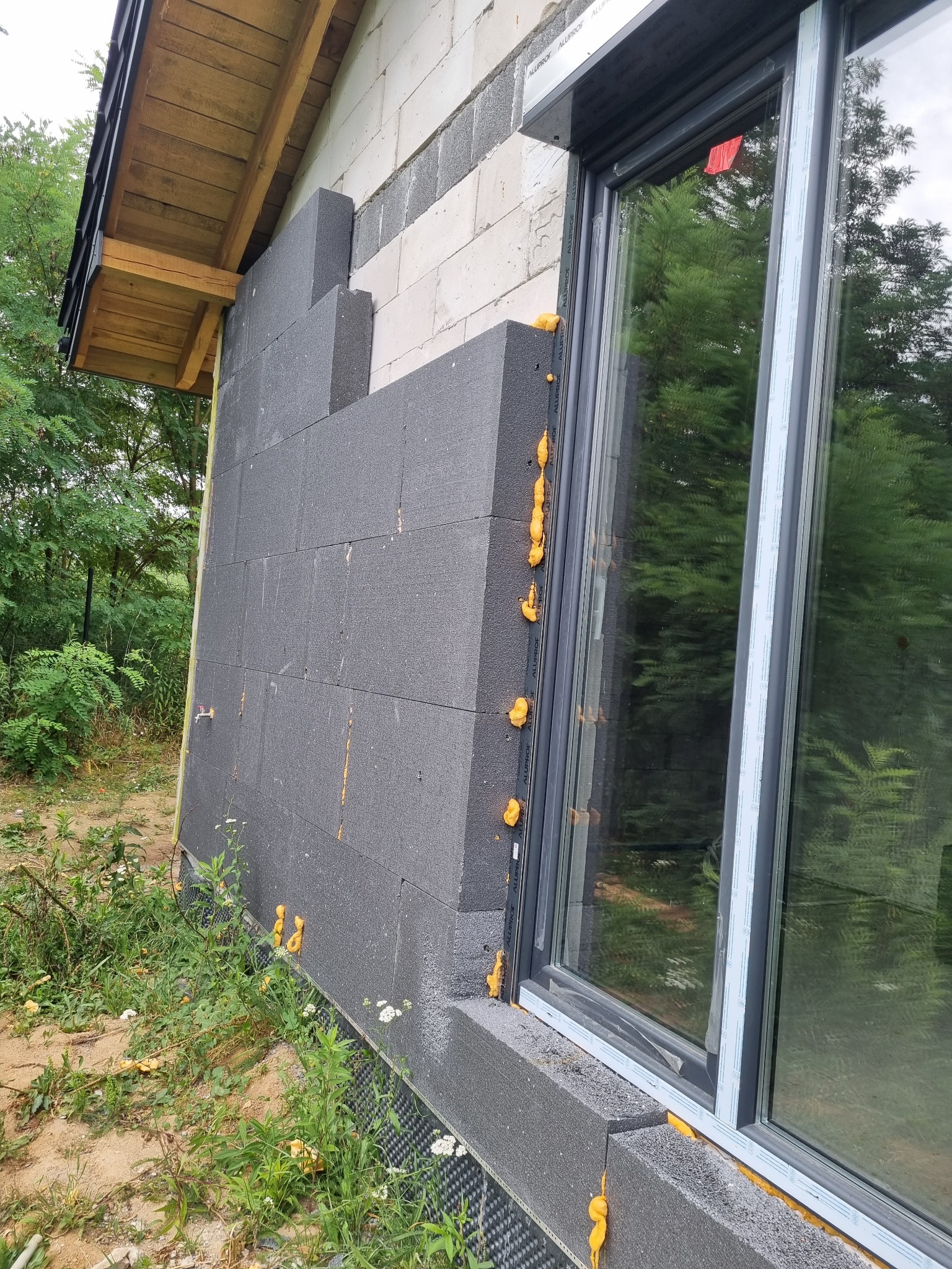 Fragment ściany domu w trakcie ocieplania styropianem grafitowym, widoczna piana montażowa wokół okna z roletą zewnętrzną, niezabezpieczone styropianem fragmenty ściany z bloczków.