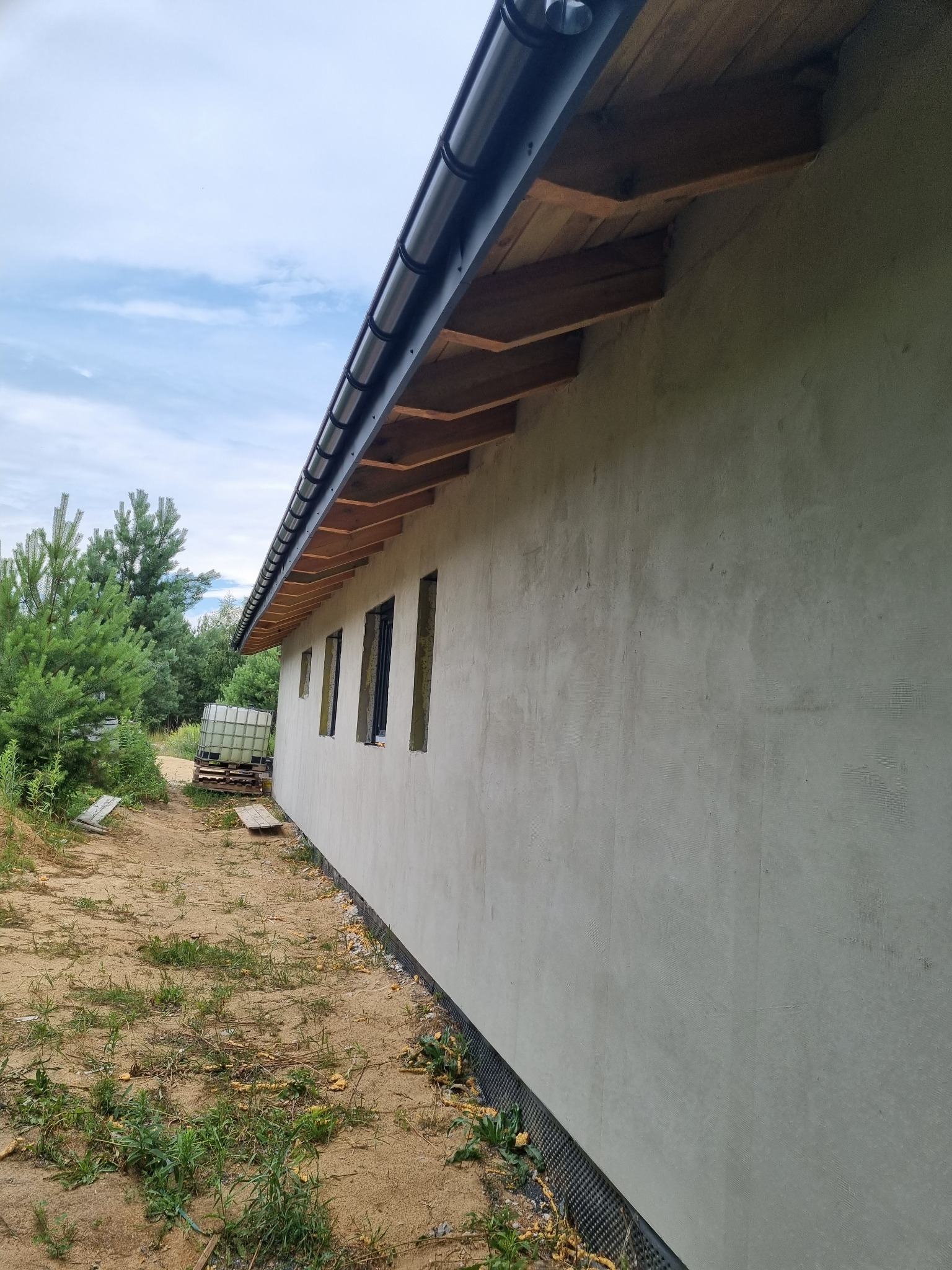 Świeżo otynkowana ściana budynku z widoczną siatką zbrojącą, czarną rynną i fragmentami drewnianej konstrukcji dachu, z oknami w trakcie montażu, na tle piaszczystego podłoża i zieleni.