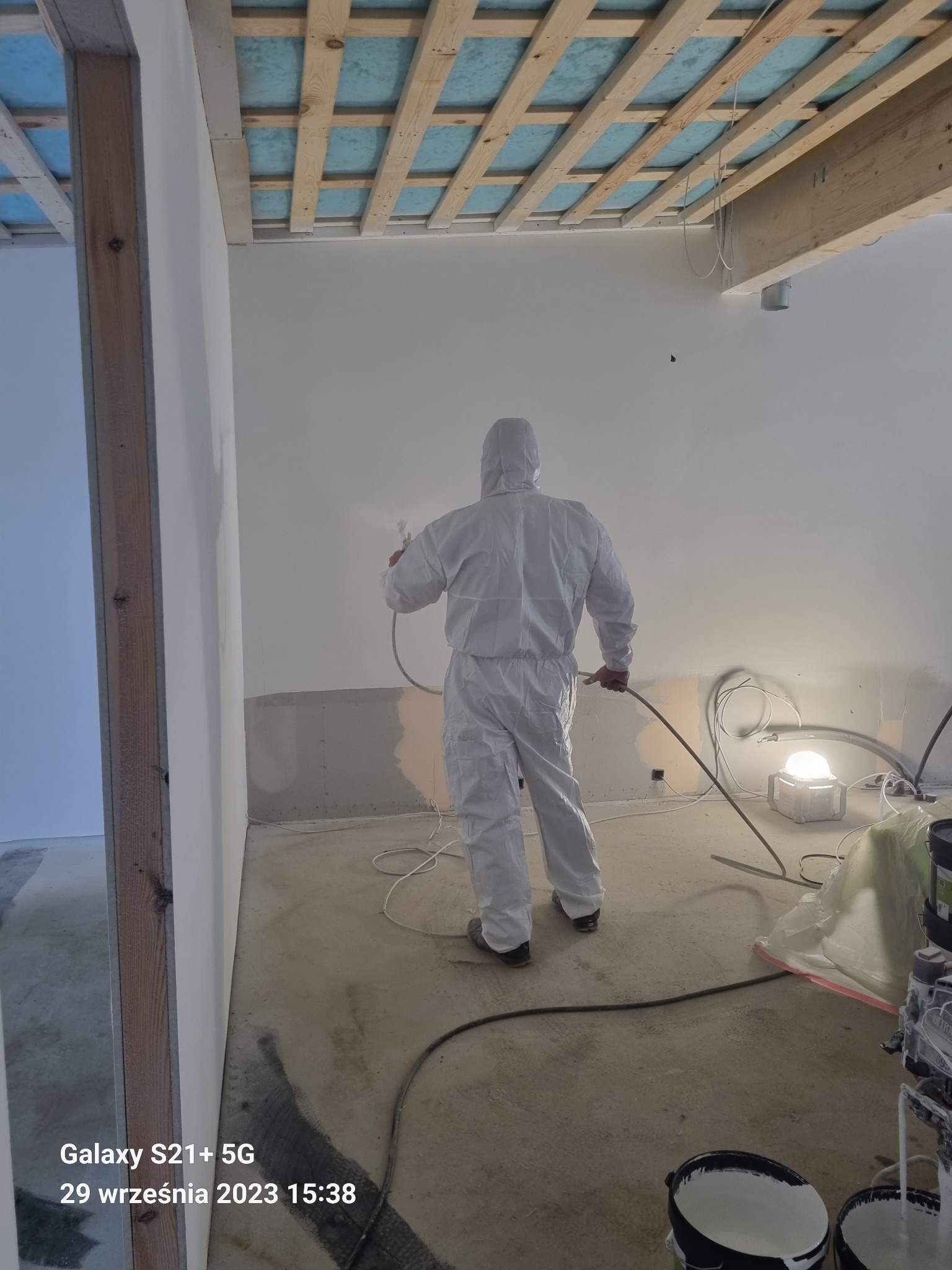 Pracownik w kombinezonie ochronnym maluje ściany wewnątrz budynku za pomocą pistoletu natryskowego, widoczne surowe wykończenie, lampa budowlana i wiaderka z farbą.