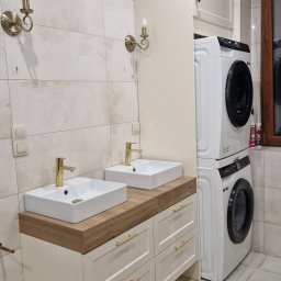 Meble na wymiar SEAL - Jasna łazienka z dwiema umywalkami nablatowymi z bateriami w kolorze złota, szafką z szufladami pod blatem z drewnopodobnym wykończeniem, pralką i suszarką w zabudowie oraz dekoracyjnymi kinkietami.