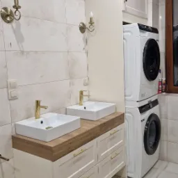 Jasna łazienka z dwiema umywalkami nablatowymi z bateriami w kolorze złota, szafką z szufladami pod blatem z drewnopodobnym wykończeniem, pralką i suszarką w zabudowie oraz dekoracyjnymi kinkietami.