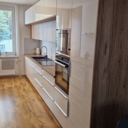 Meble na wymiar SEAL - Nowoczesna kuchnia na wymiar z jasnymi, lakierowanymi frontami szafek i blatem z drewna, zintegrowanym piekarnikiem i mikrofalówką, widok z perspektywy stołu jadalnego.