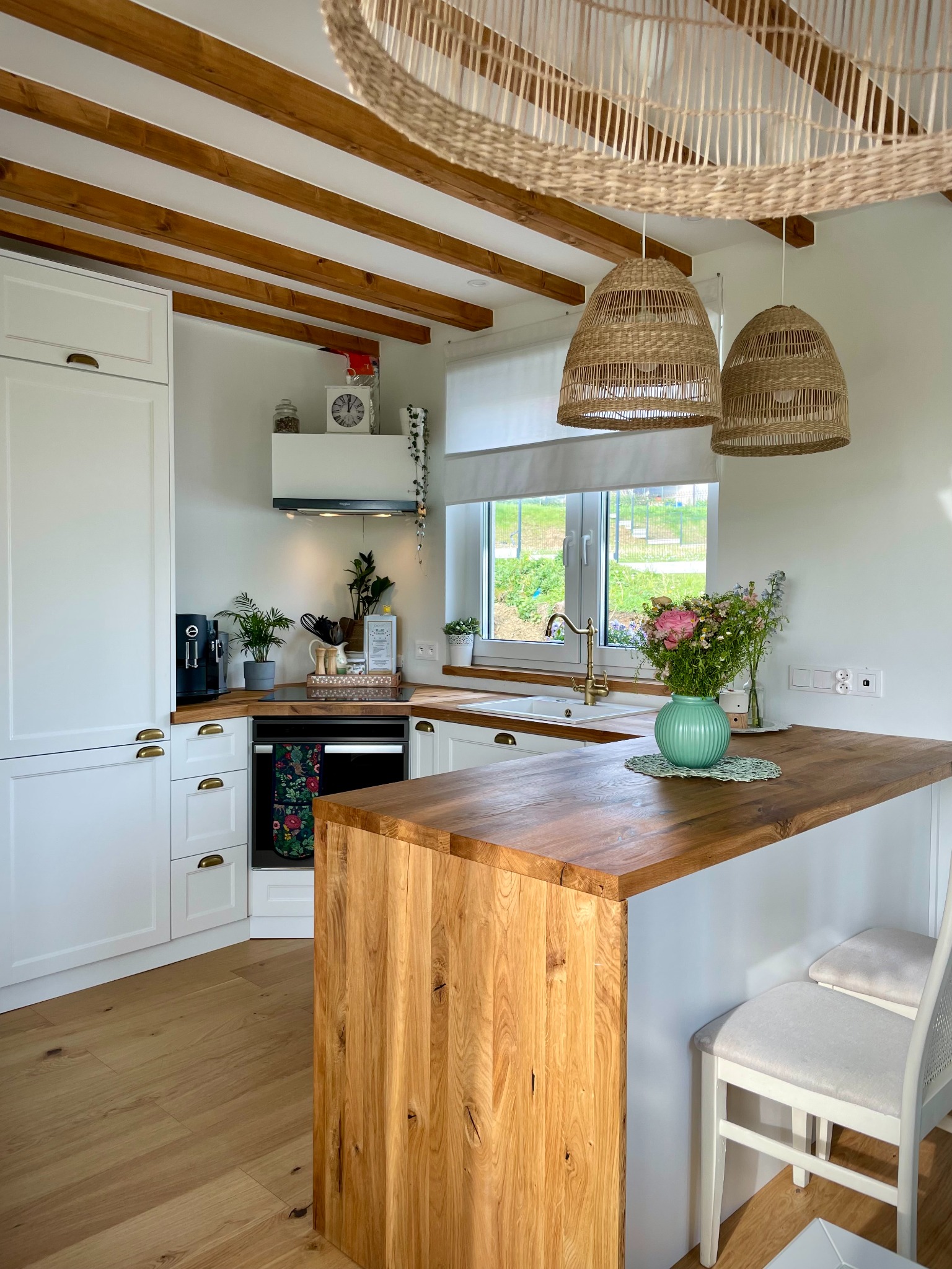 Jasna kuchnia z drewnianym blatem wyspowym, białymi szafkami z mosiężnymi uchwytami, drewnianymi belkami stropowymi i wiklinowymi lampami, bukiet kwiatów w wazonie na blacie.