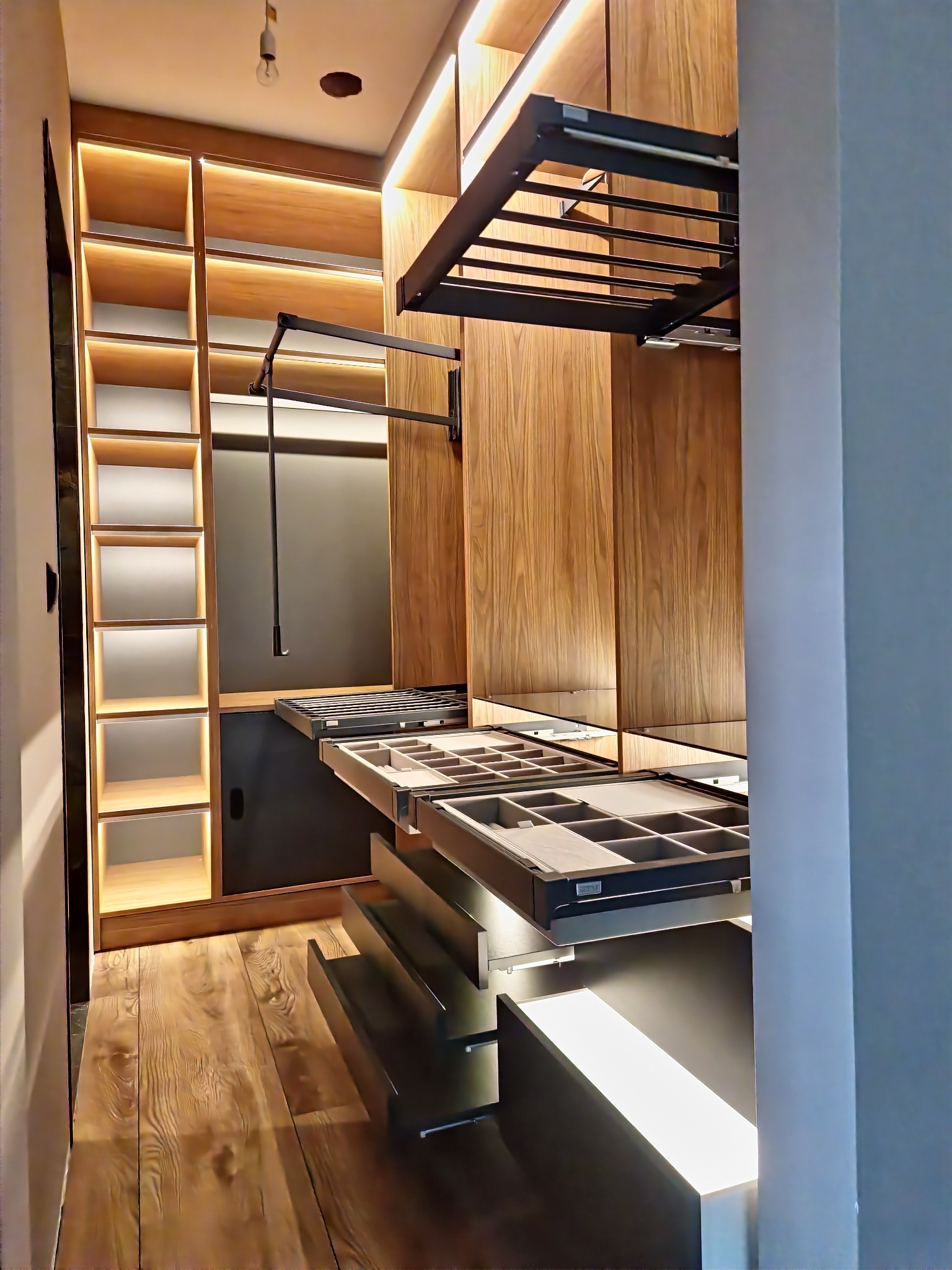 Wnętrze garderoby z drewnianymi elementami, oświetleniem LED i wysuwanymi szufladami na akcesoria, widok z perspektywy wejścia.