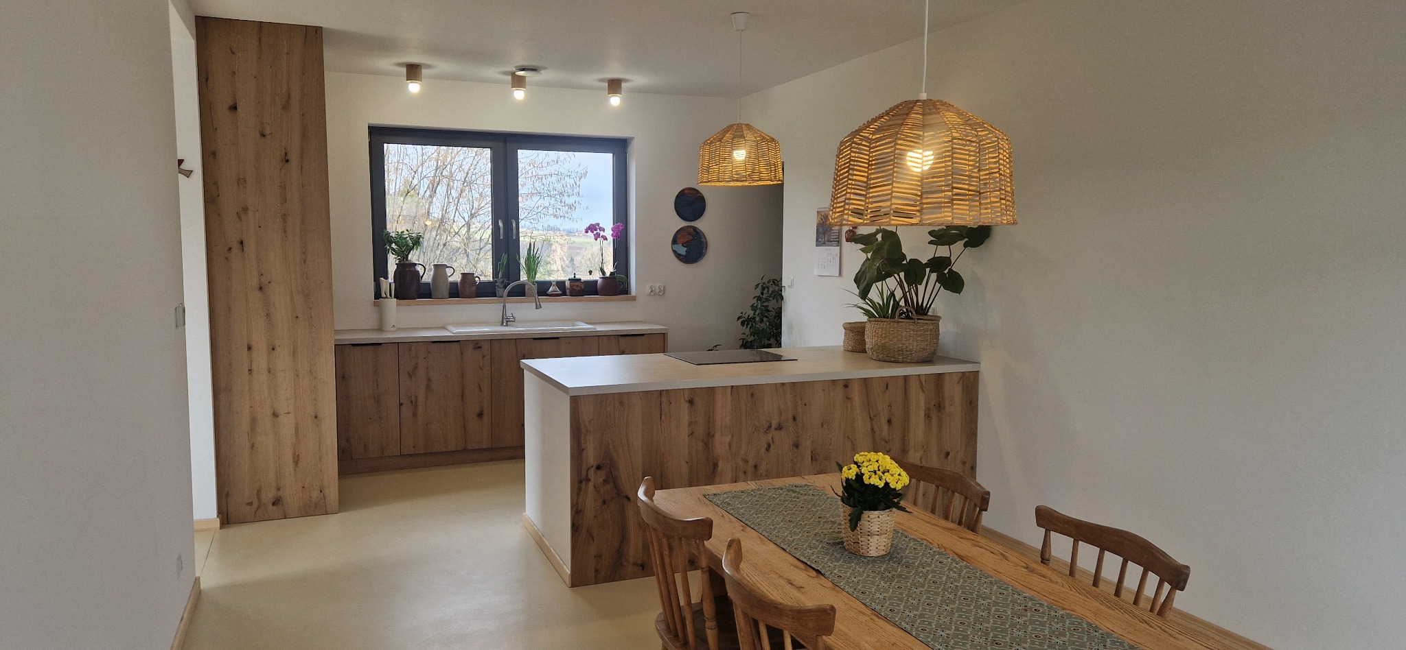 Jasna kuchnia w Ustroniu z drewnianymi frontami, wyspą i rattanowymi lampami, widok na stół z żółtymi kwiatami.