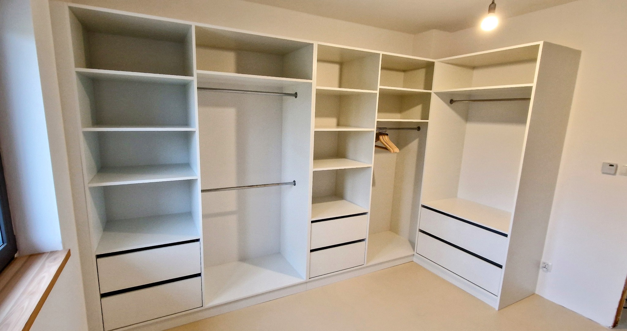 Wnętrze garderoby z białymi szafami wnękowymi z szufladami z czarnymi uchwytami i półkami, widoczny wieszak na ubrania.