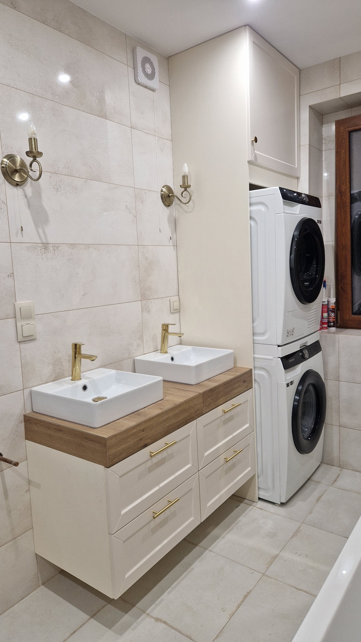 Jasna łazienka z dwiema umywalkami nablatowymi z bateriami w kolorze złota, szafką z szufladami pod blatem z drewnopodobnym wykończeniem, pralką i suszarką w zabudowie oraz dekoracyjnymi kinkietami.