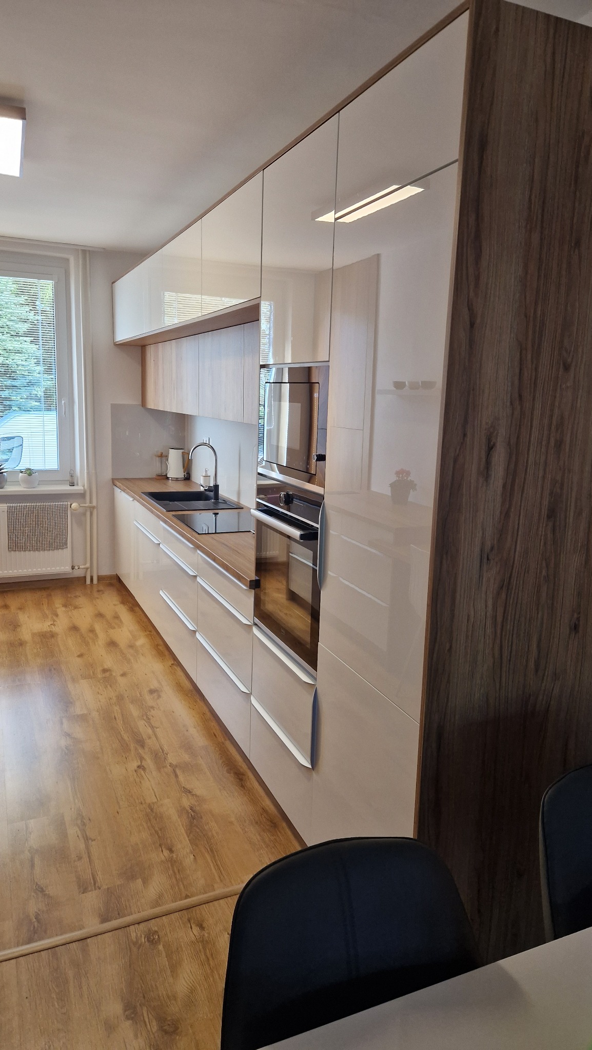 Nowoczesna kuchnia na wymiar z jasnymi, lakierowanymi frontami szafek i blatem z drewna, zintegrowanym piekarnikiem i mikrofalówką, widok z perspektywy stołu jadalnego.