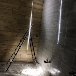 Remont łazienki w trakcie: położone płytki imitujące drewno na jednej ścianie, szare płytki na drugiej, drabina oświetlona lampą LED, podłoga zabezpieczona folią malarską.