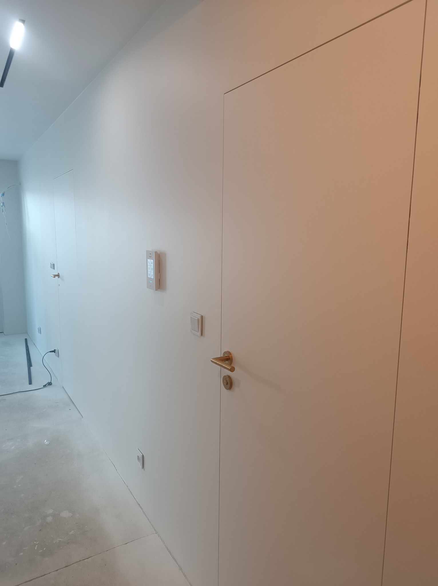 Minimalistyczne wnętrze: białe ściany, ukryte drzwi z złotą klamką, dotykowy panel sterowania i gładka podłoga w trakcie wykańczania.