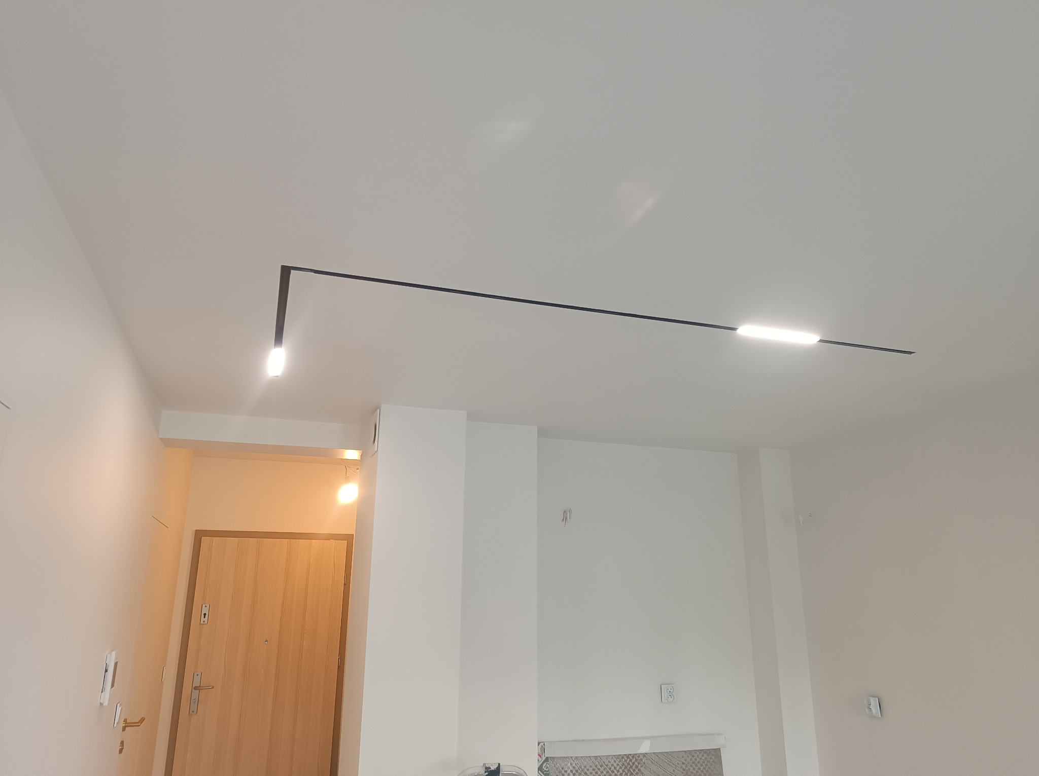 Nowoczesna lampa sufitowa w kształcie litery L z dwoma punktami świetlnymi w minimalistycznym, białym wnętrzu mieszkania po remoncie.