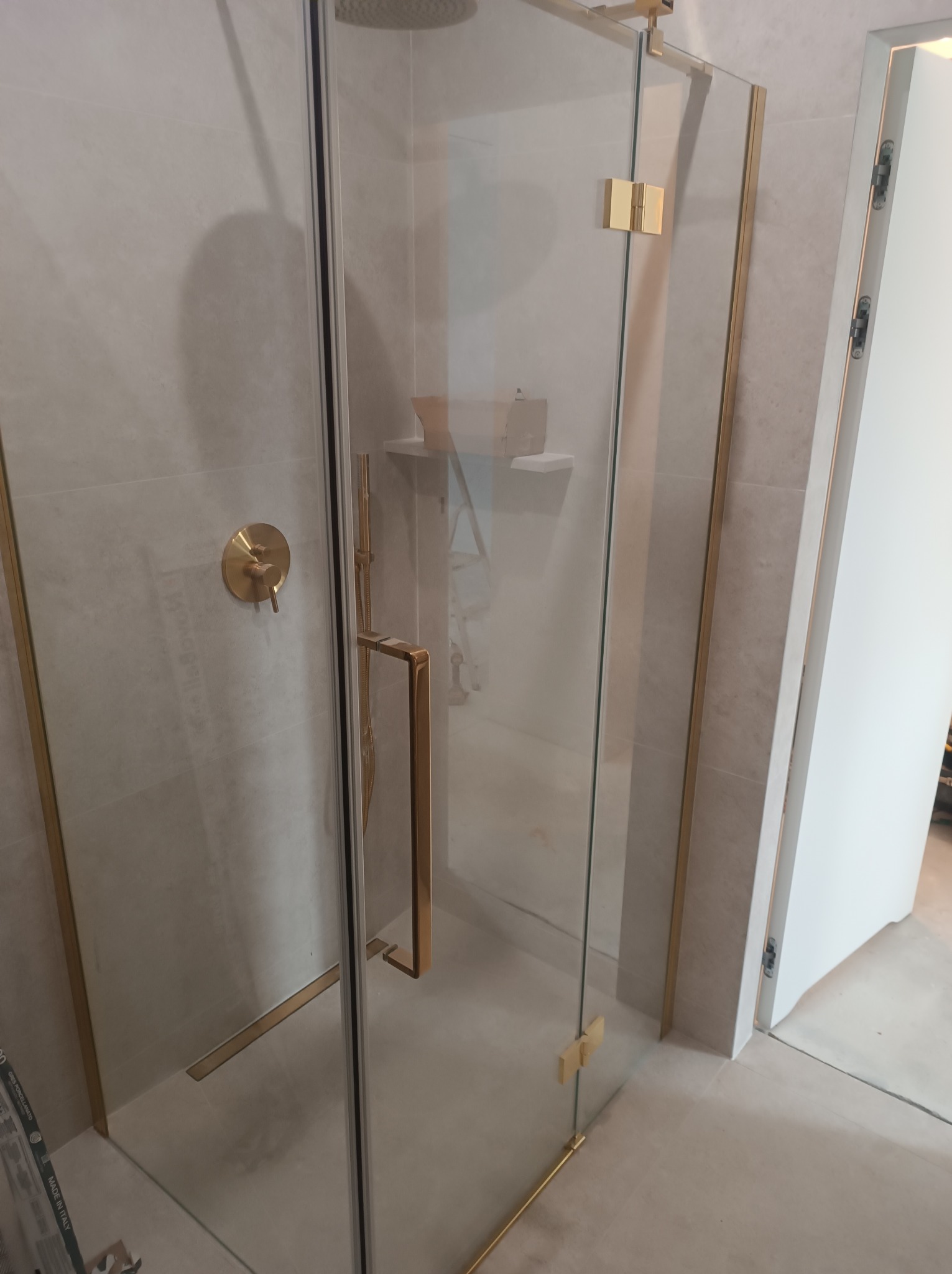 Szklana kabina prysznicowa z złotymi okuciami i baterią w odcieniach beżu i bieli, widoczne płytki na ścianach i podłodze.