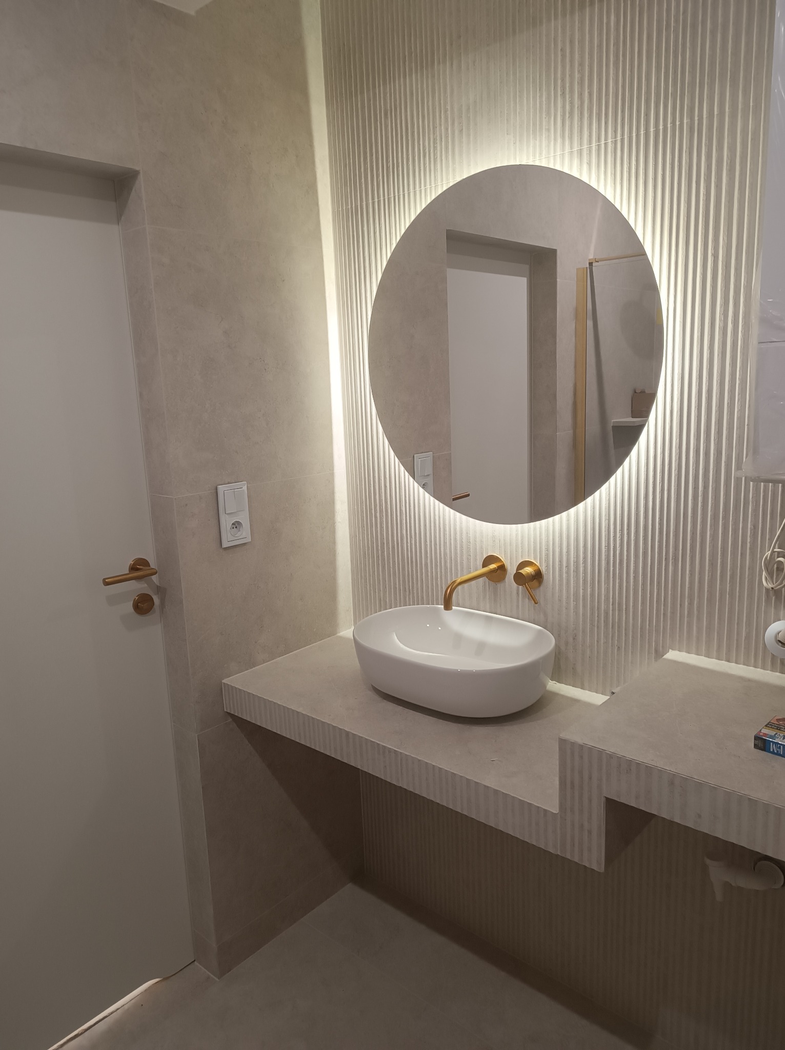 Nowoczesna łazienka z okrągłym lustrem podświetlanym LED, białą umywalką nablatową i złotą armaturą na blacie z efektem ryflowania.