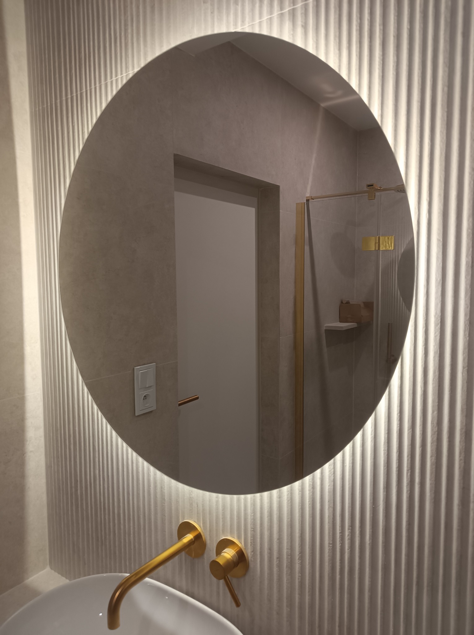 Nowoczesna łazienka z okrągłym lustrem podświetlanym LED, złotą armaturą i płytkami z delikatnym wzorem pionowych pasków, widoczny fragment kabiny prysznicowej z elementami w kolorze złota.