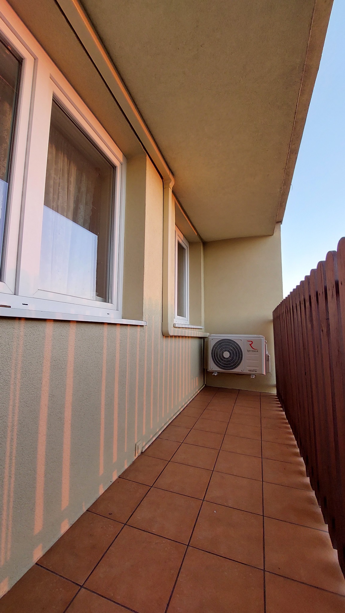 Zewnętrzna jednostka klimatyzacji Rotenso umieszczona na balkonie, z widocznymi promieniami zachodzącego słońca na ścianie budynku i brązowymi płytkami na podłodze.