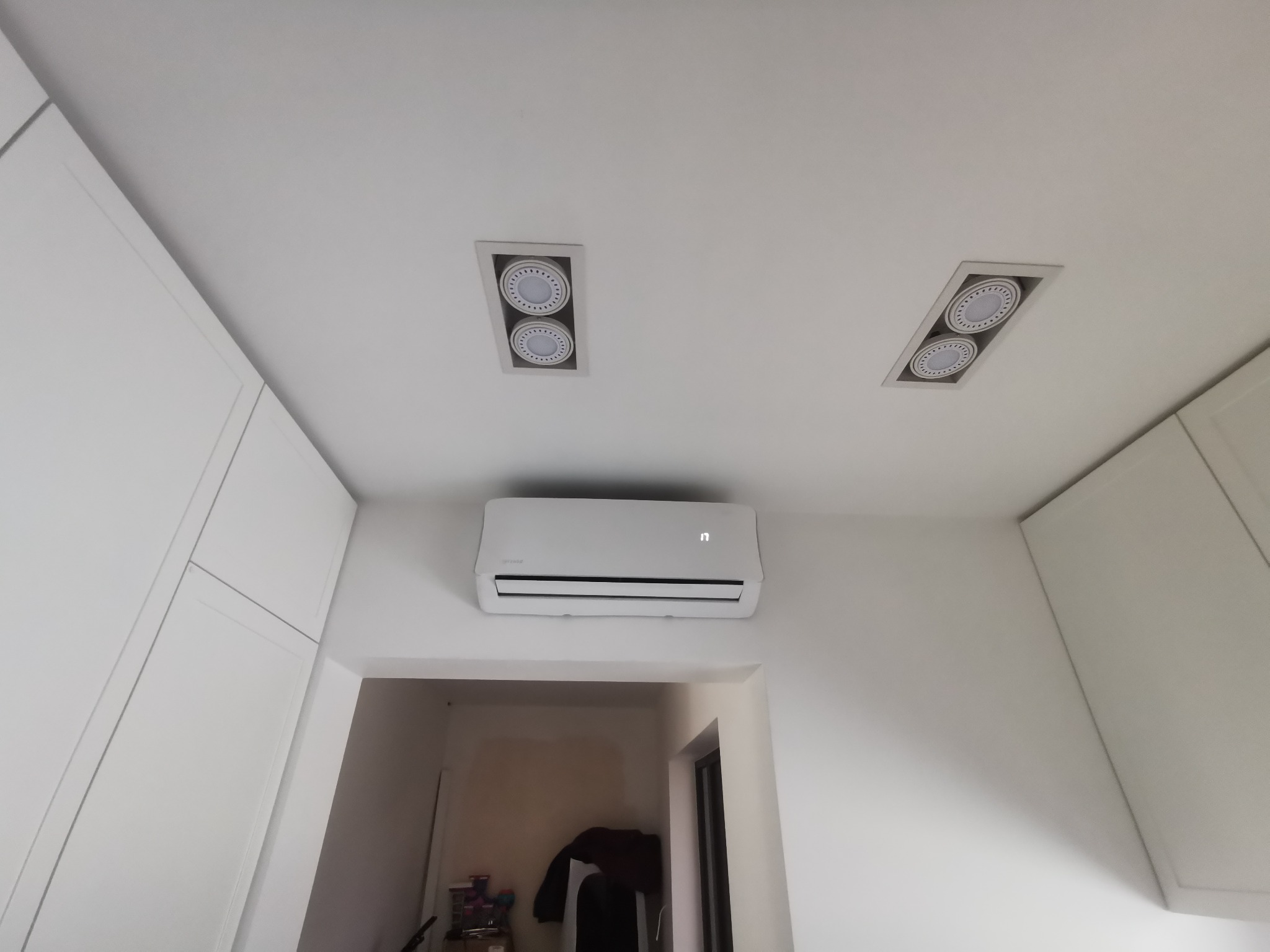 Biały klimatyzator ścienny zamontowany w jasnym pomieszczeniu z białymi szafkami i oświetleniem punktowym w suficie.