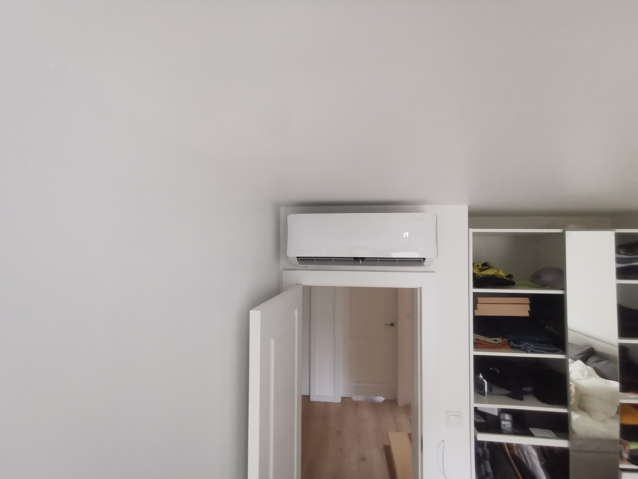 Biały klimatyzator ścienny zamontowany nad otwartymi drzwiami w minimalistycznym wnętrzu z szafą wnękową po prawej stronie.