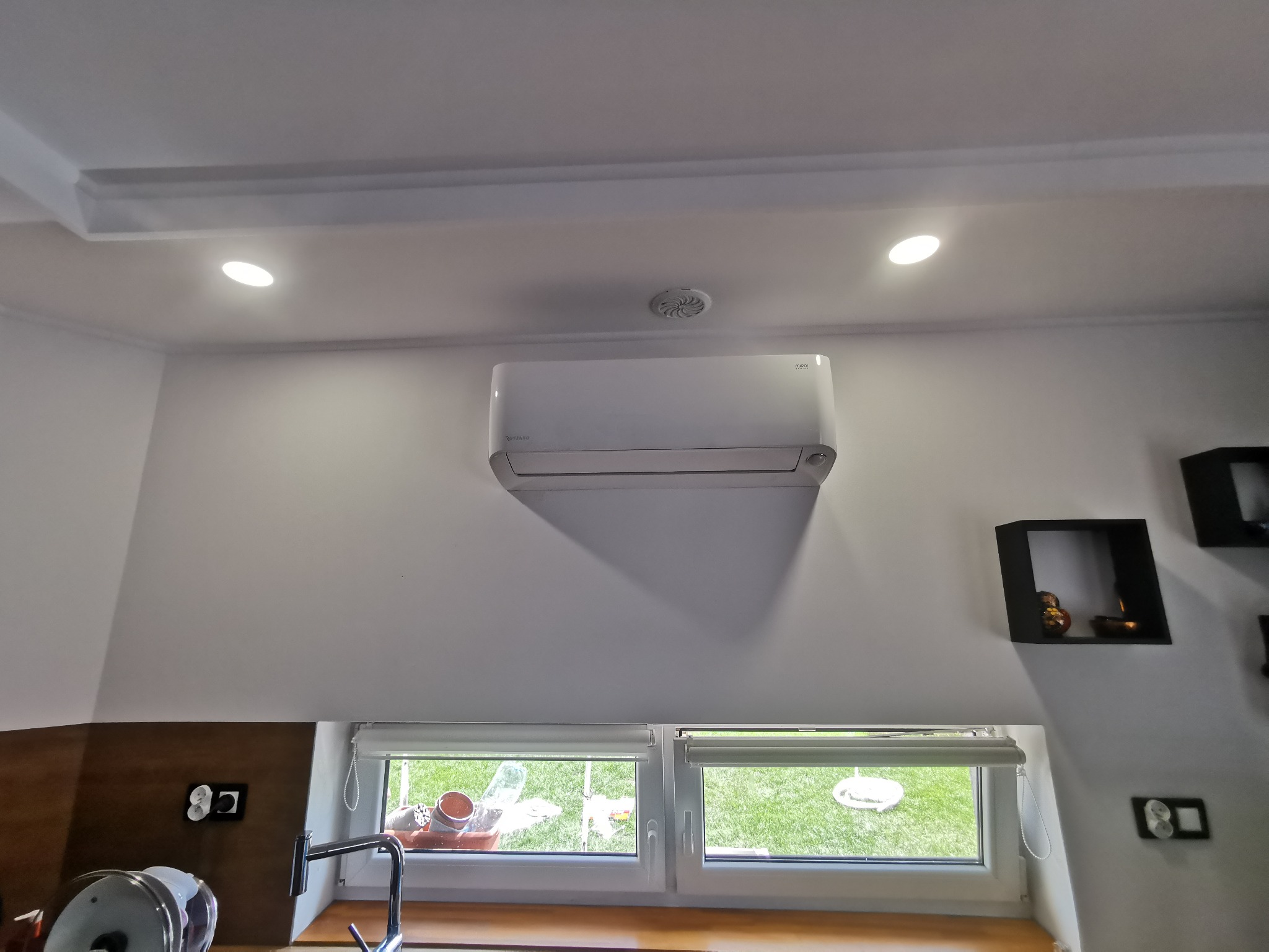 Biały klimatyzator ścienny zamontowany nad oknem w jasnym pomieszczeniu, widoczne elementy wystroju wnętrza i fragment blatu kuchennego z kranem.