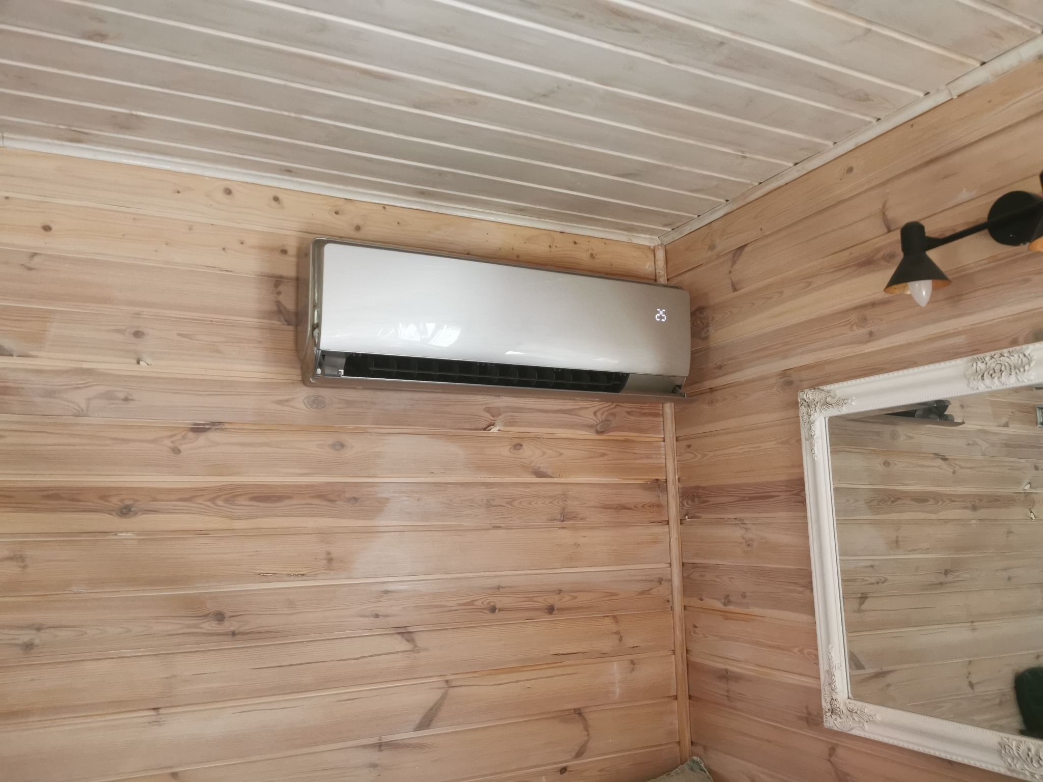Biały klimatyzator zamontowany na drewnianej ścianie w domu, wyświetlający temperaturę 25 stopni Celsjusza, widoczny fragment lustra w ozdobnej ramie i lampka ścienna.