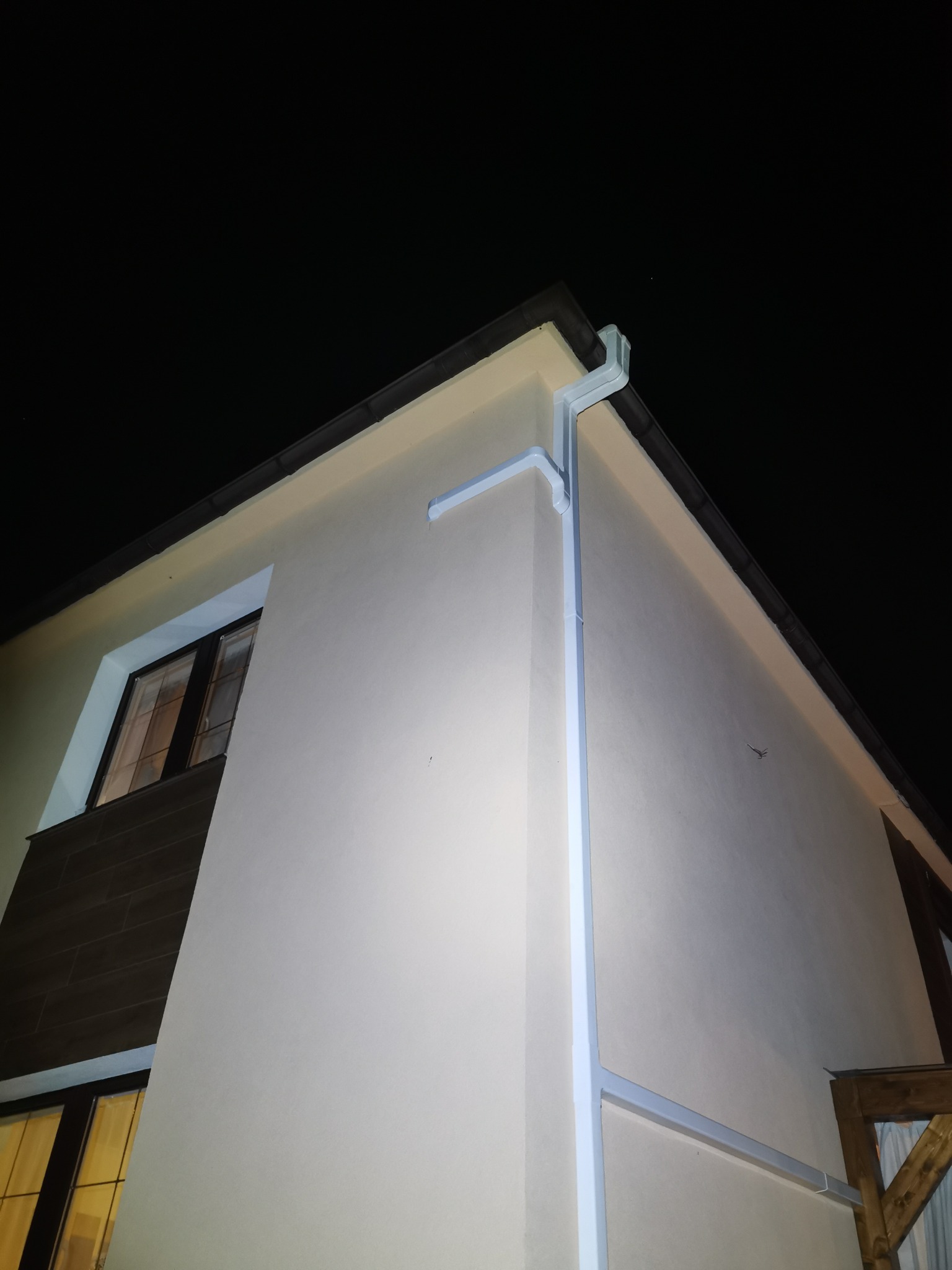 Nocne ujęcie narożnika domu z zamontowanym, jasnym systemem orynnowania, widoczne okno z podświetlonym wnętrzem.