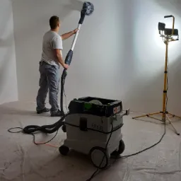 Pracownik w szarych spodniach i białej koszulce szlifuje ścianę za pomocą szlifierki z długim uchwytem podłączonej do odkurzacza przemysłowego, oświetlonej halogenem na statywie, na podłodze folia...