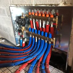 HydroDave - Rozdzielacz instalacji ogrzewania podłogowego z czerwono-niebieskimi rurami w izolacji, zamontowany na ścianie z cegły, widoczne regulatory przepływu.