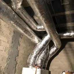 Srebrne, izolowane rury wentylacyjne podwieszone pod sufitem, podłączone do białej jednostki centralnej, widoczne na tle surowej ściany z cegły.