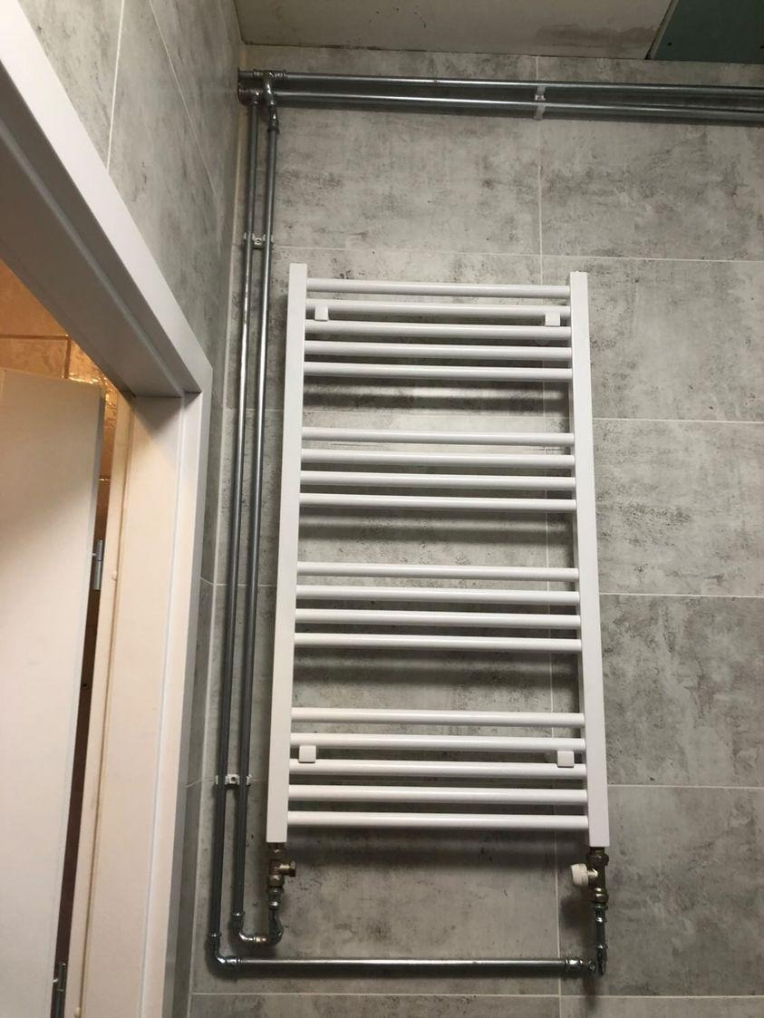 Biały grzejnik łazienkowy drabinkowy zamontowany na szarej, ceramicznej ścianie, z widocznymi chromowanymi rurami doprowadzającymi wodę i zaworami regulacyjnymi.