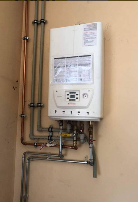 Biały piec gazowy zamontowany na ścianie z beżową farbą, widoczne podłączenia rur miedzianych i stalowych, zawory oraz panel sterowania z wyświetlaczem.