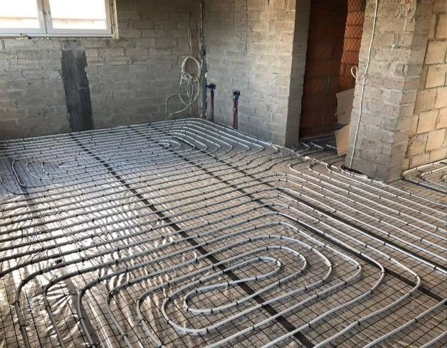 Rozłożone rury systemu grzewczego na folii izolacyjnej, przygotowane do zalania wylewką w surowym wnętrzu z ceglanymi ścianami.