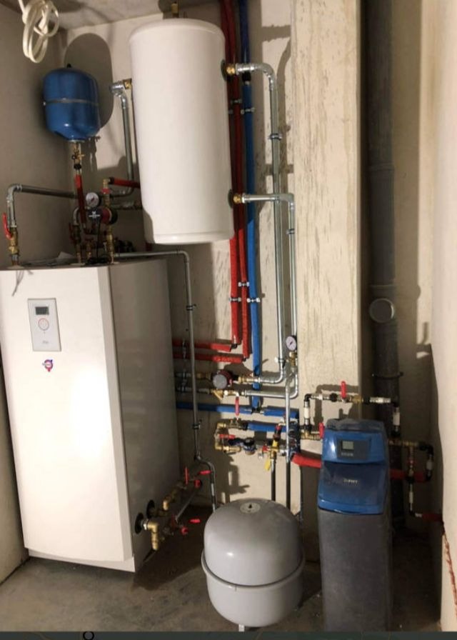 Kompletna instalacja grzewcza w piwnicy z piecem, zbiornikiem buforowym, naczyniem przeponowym i zmiękczaczem wody, rury miedziane i plastikowe w kolorze czerwonym i niebieskim, szare ściany...