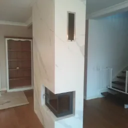 Biały, marmurkowy kominek narożny z czarnym wkładem, wkomponowany w ścianę działową, z widoczną kratką wentylacyjną i fragmentem drewnianej podłogi.