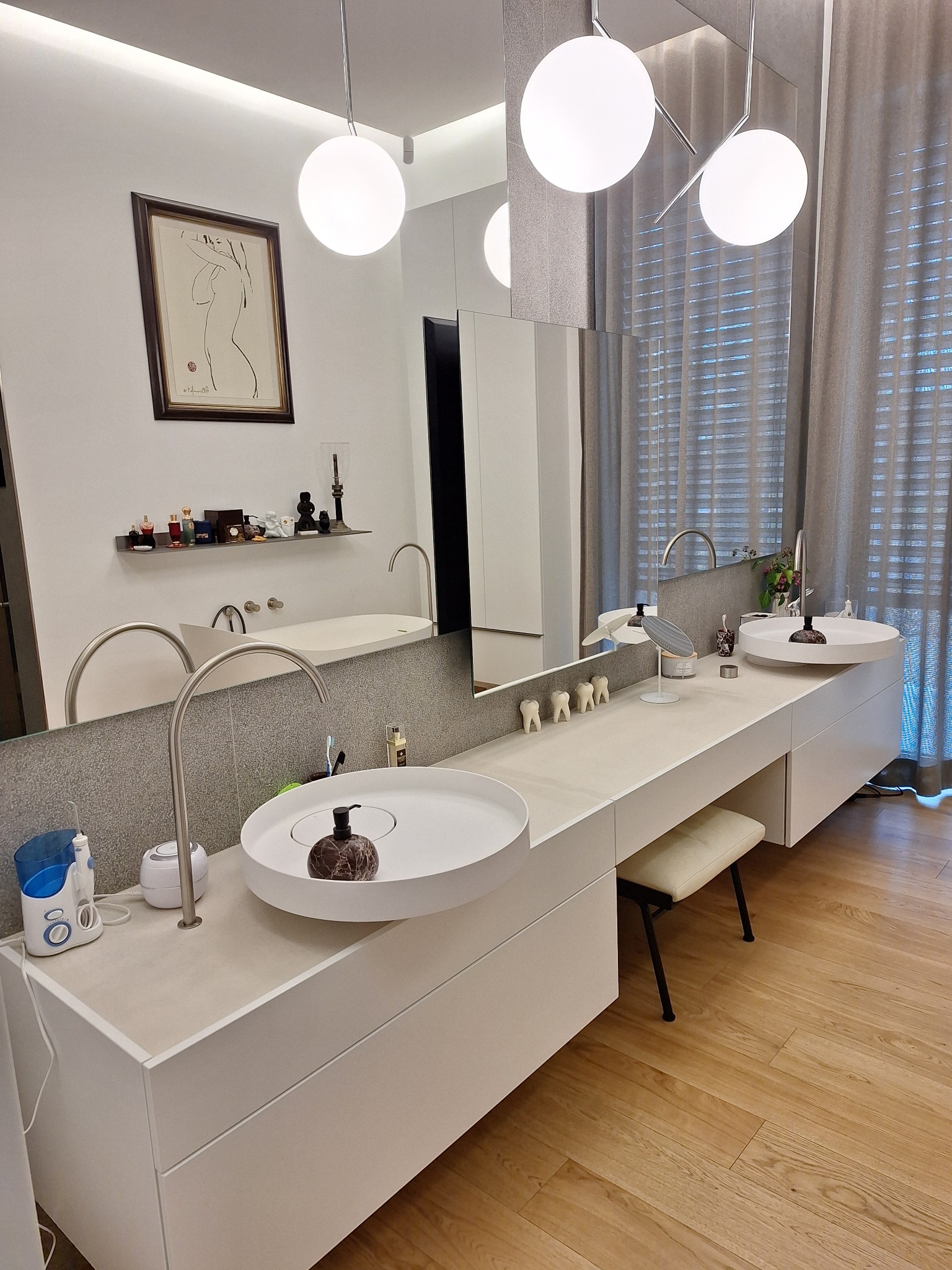 Nowoczesna łazienka z dwoma okrągłymi umywalkami na blacie, podświetlane lustro, minimalistyczne lampy i drewniana podłoga. W tle zasłonięte okno.