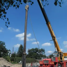 Żuraw budowlany podnosi metalową kolumnę z otworami na placu budowy, w tle drzewa i błękitne niebo z chmurami.