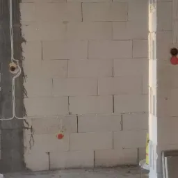 Świeżo położona instalacja elektryczna w surowym wnętrzu z białych bloczków, widoczne kable i puszki podtynkowe, okno z widokiem na zieleń w tle, na podłodze widoczne wiadra po farbie.