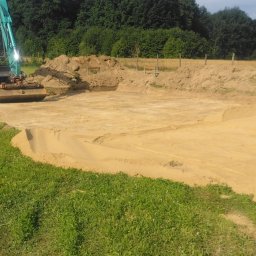 FibroForm płyty fundamentowe - Turkusowa koparka kołowa przy wykopie pod fundamenty, z płaskim dnem i usypanymi hałdami piasku, na tle zielonych drzew.