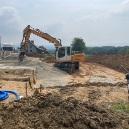 FibroForm płyty fundamentowe - Żółto-biała koparka Liebherr 900 na placu budowy, przygotowująca teren pod fundamenty. Widoczna niebieska rura drenażowa i sprzęt geodezyjny, w tle wiejskie zabudowania.
